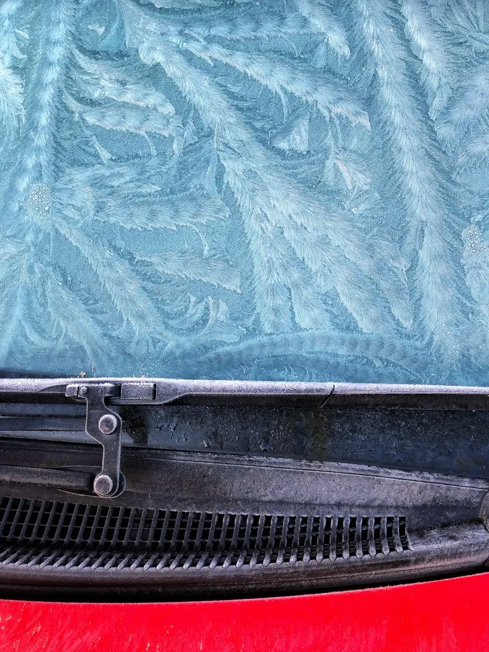IJskristallen op een autoruit boven een zwarte ruitenwisser en een rode carrosserie