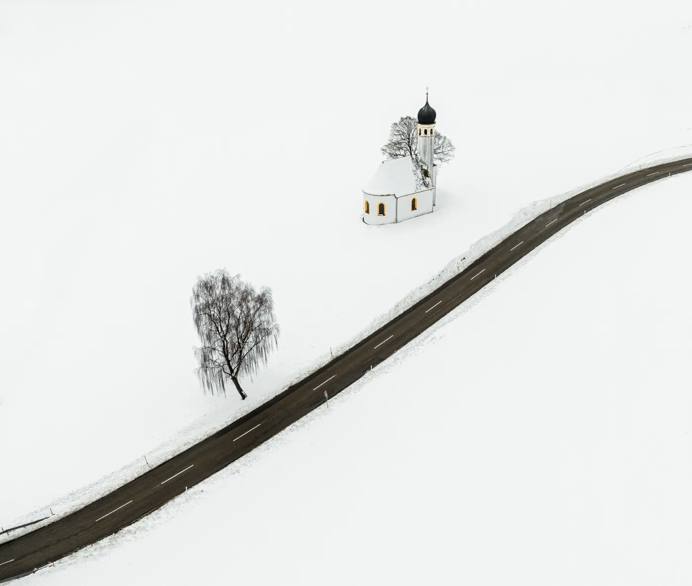 Luchtfoto van een klein wit kerkje naast een bochtige weg in een besneeuwd landschap.