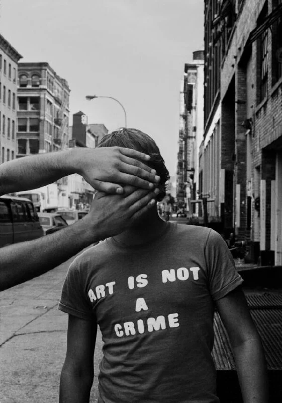Zwart-witfoto van een jongen in een straat met het T-shirt ‘ART IS NOT A CRIME’, terwijl twee handen zijn gezicht bedekken.