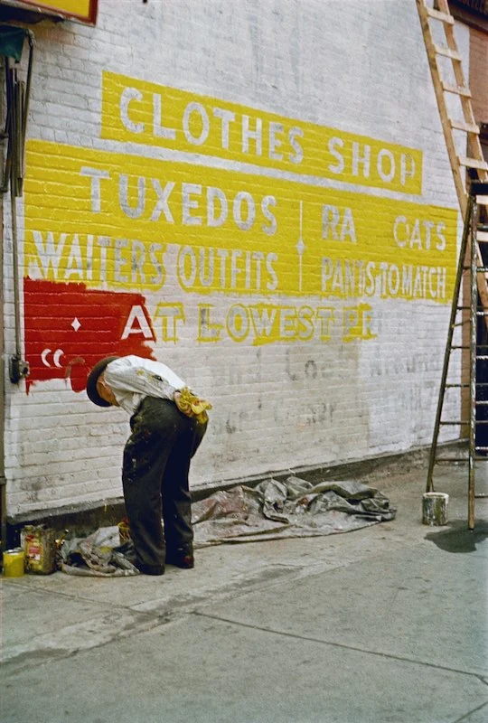 Een schilder werkt aan een gele en rode reclameschildering op een bakstenen muur, gefotografeerd door Saul Leiter.