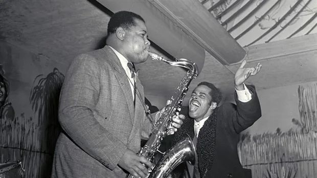 Zwart-witfoto van een saxofonist en een man die meebeweegt tijdens een jazzoptreden.