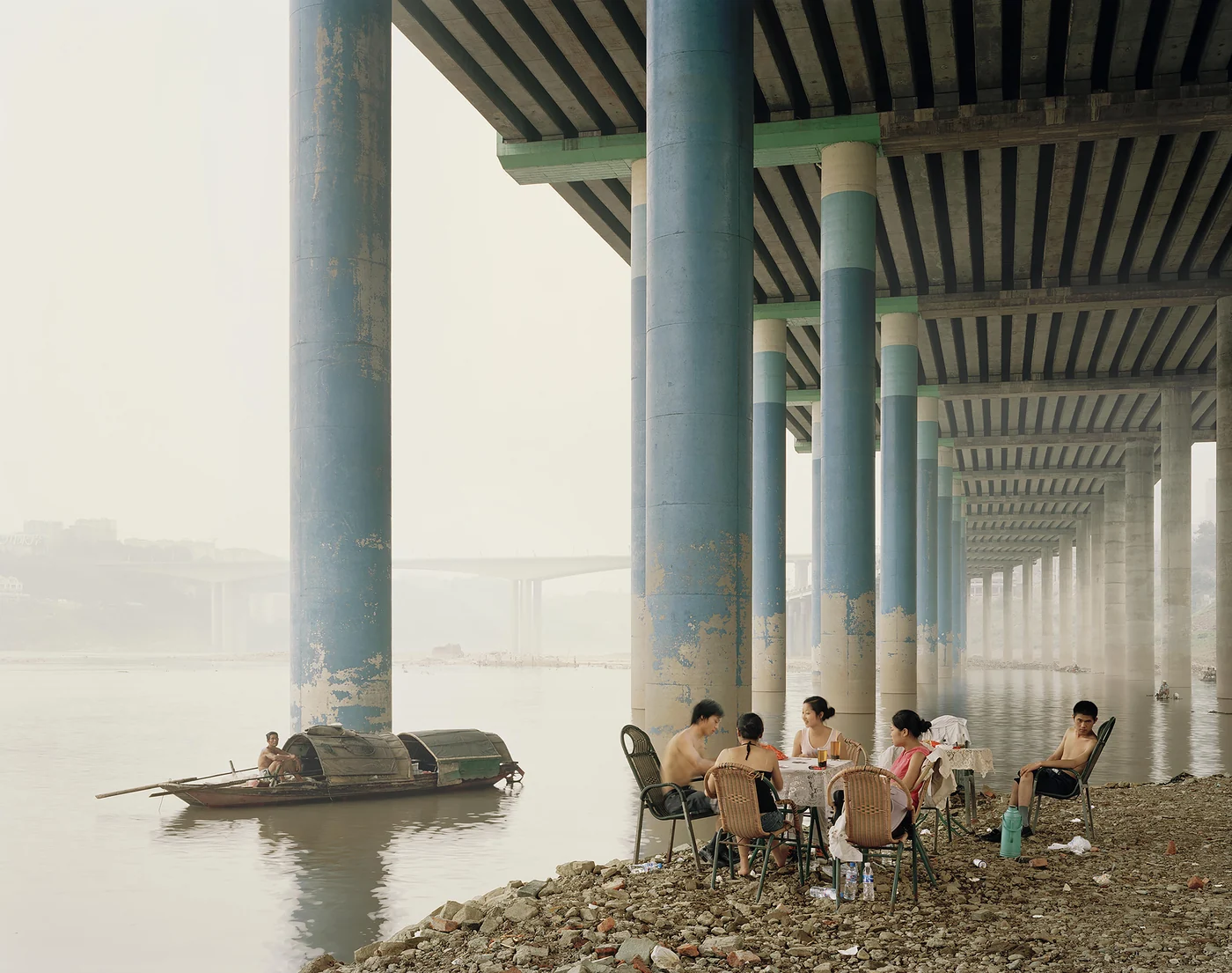 Mensen zitten aan een tafel onder een brug bij een rivier, terwijl een kleine boot voorbijvaart.