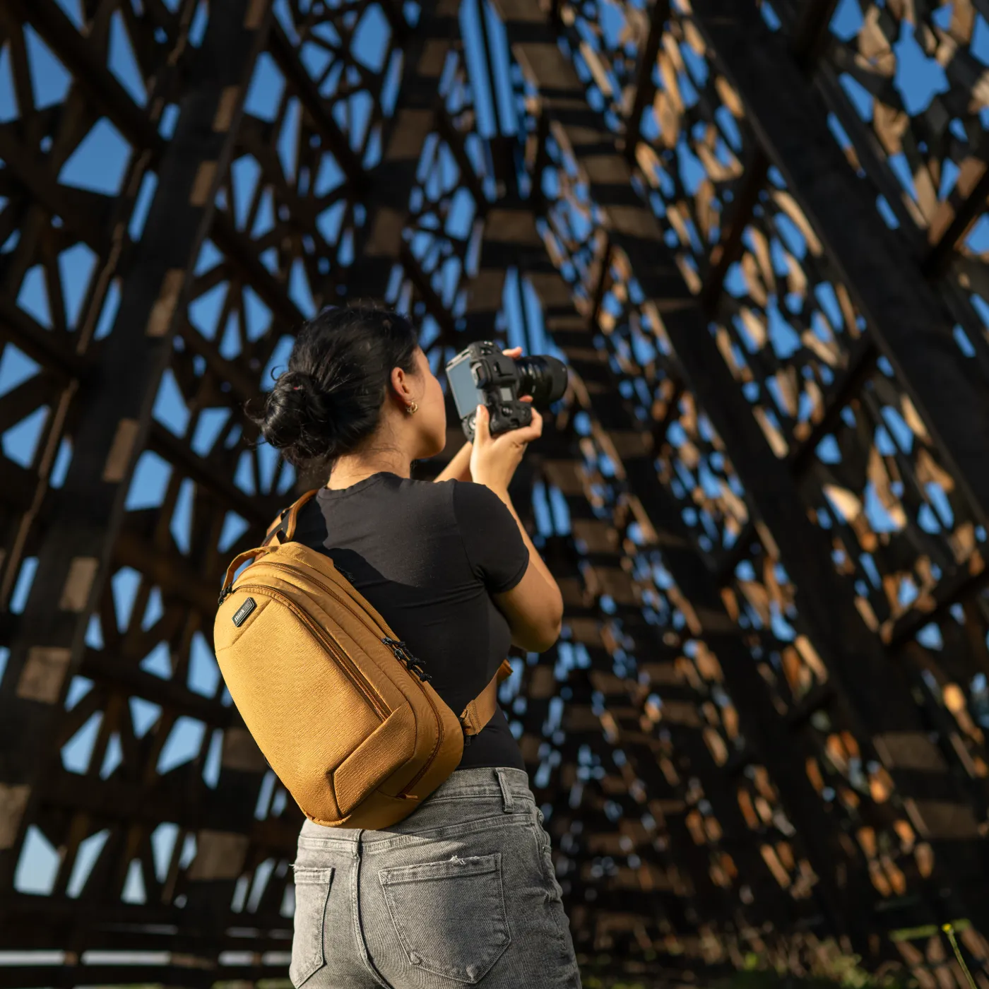 Vrouw met camera en bruine Think Tank slingtas onder een metalen constructie.