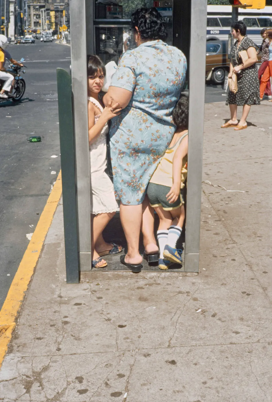 uit de tentoonstelling Helen Levitt: City at Play in Kunsthal Rotterdam: Kleurenfoto van een vrouw en twee kinderen die dicht opeen in een telefooncel aan een drukke straat in New York staan