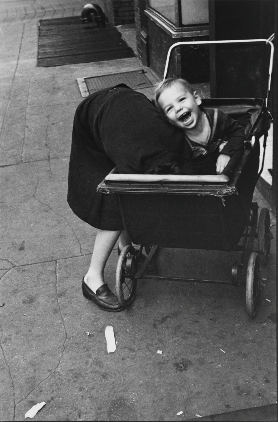 uit de tentoonstelling Helen Levitt: City at Play in Kunsthal Rotterdam: Zwart-witfoto van een lachend kind in een kinderwagen terwijl een vrouw vooroverbuigt