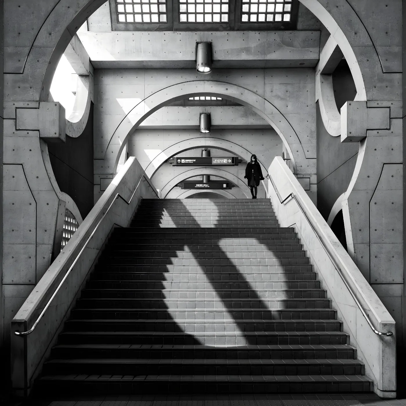 Betonnen stationshal met brede trap, ronde bogen en een persoon bovenaan de trap
