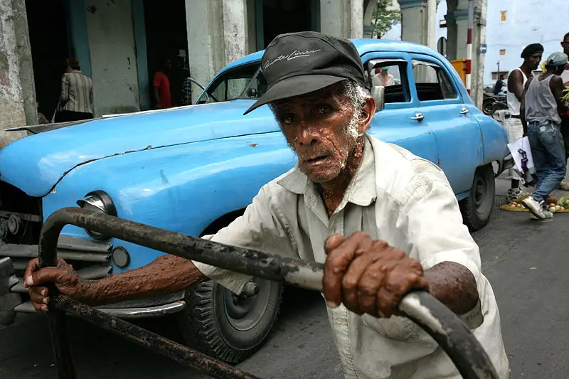 Older man in a cap holding a metal steering rod in front of a blue vintage car on the street