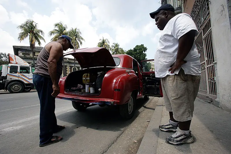 Two men standing beside a red vintage car with its trunk open on a palm-lined street