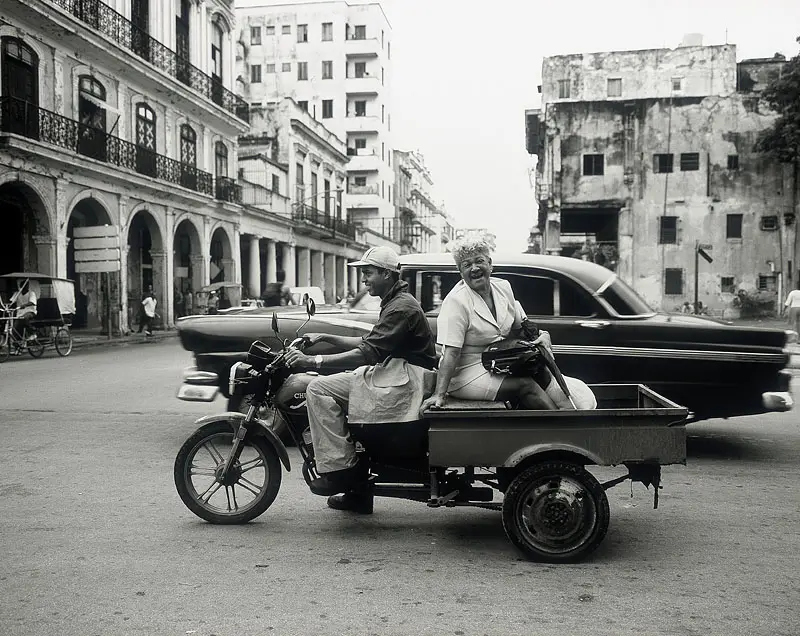 Black-and-white photograph of a woman riding in a three-wheeled motorized cargo bike on the street