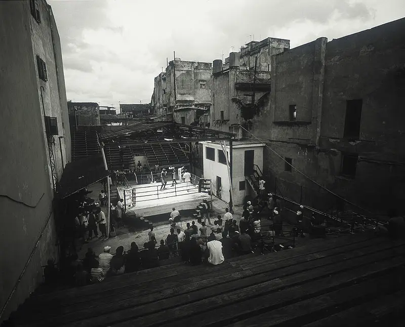 Black-and-white photograph of a boxing ring with spectators between tall buildings