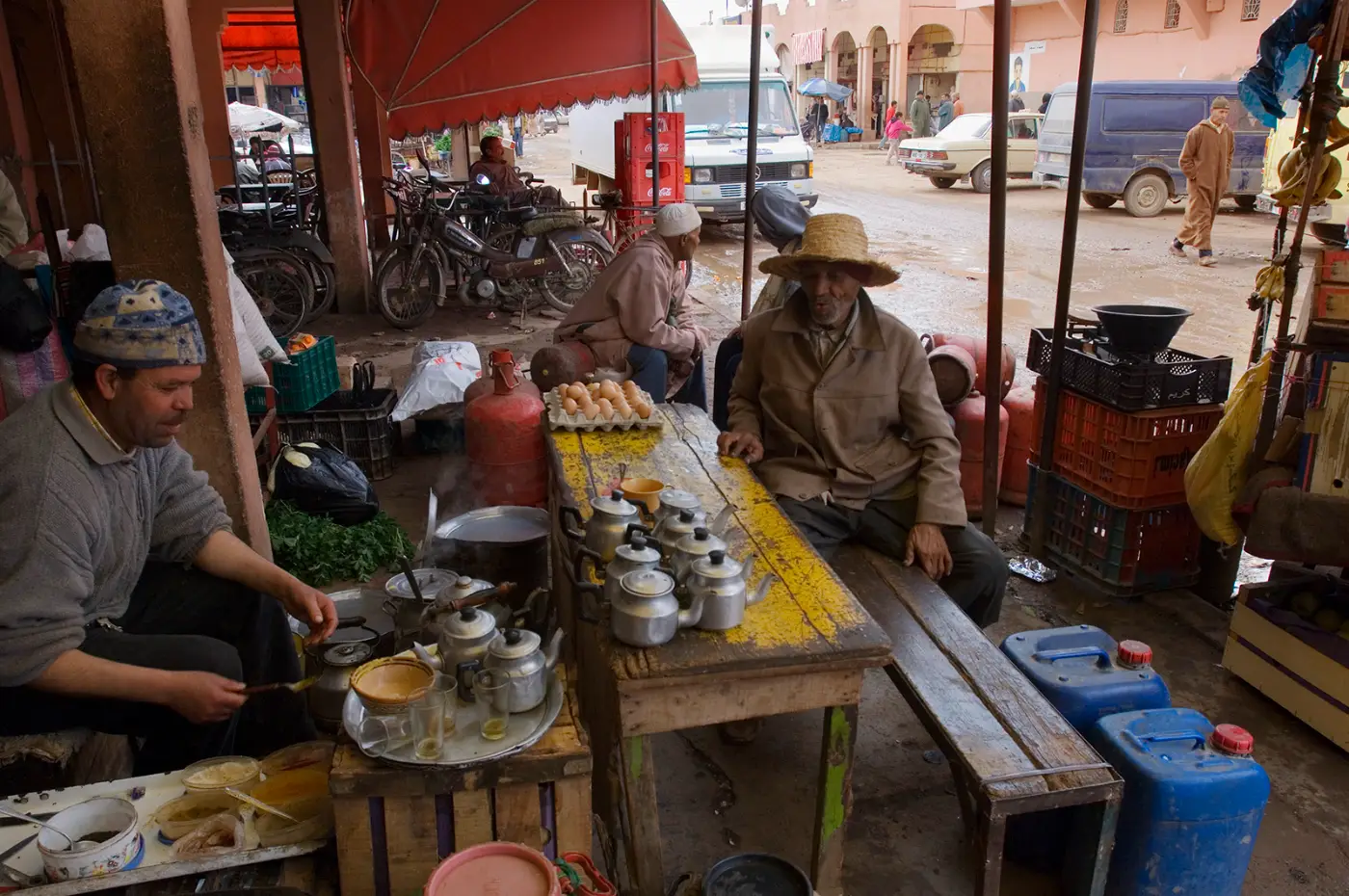 Twee mannen zitten aan een lage tafel met theepotten en eieren in een open marktkraam