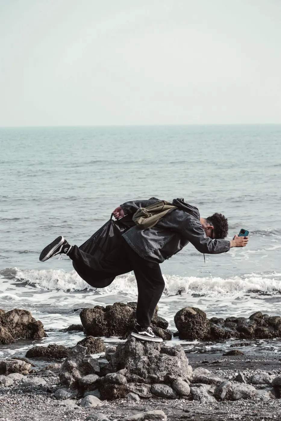 Man fotografeert met smartphone terwijl hij bijna zijn evenwicht verliest op rotsen aan zee