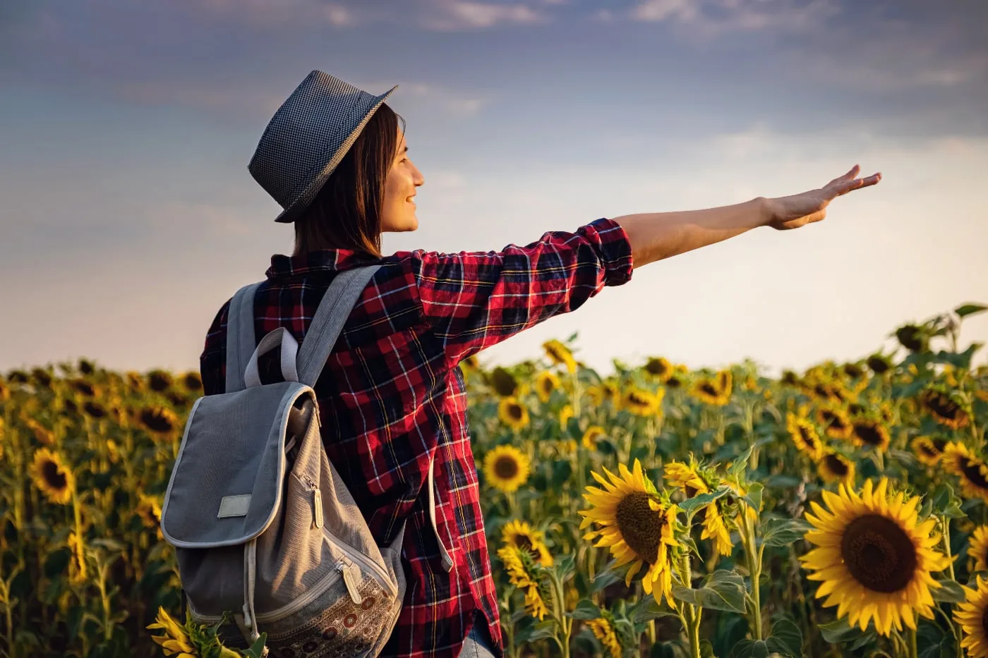 Woman with a backpack and hat in a sunflower field at sunset.