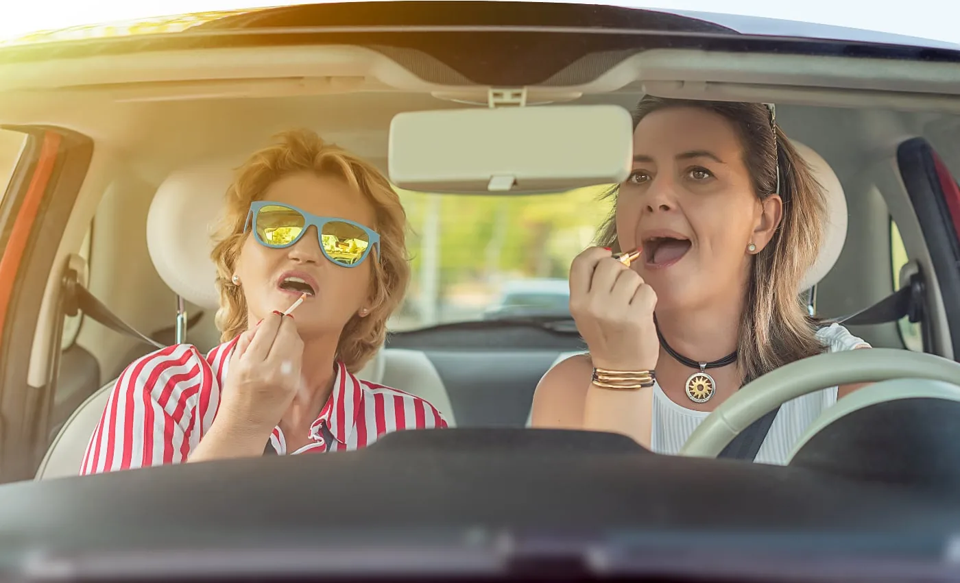 Two women apply lipstick in a sunlit car