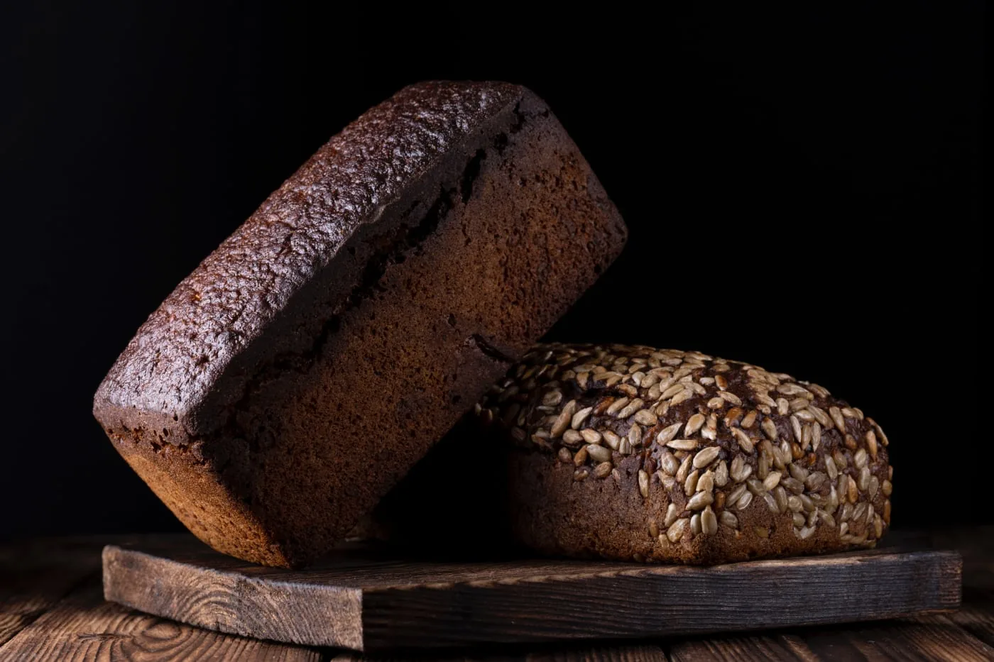 Two dark loaves of bread on a wooden board against a black background