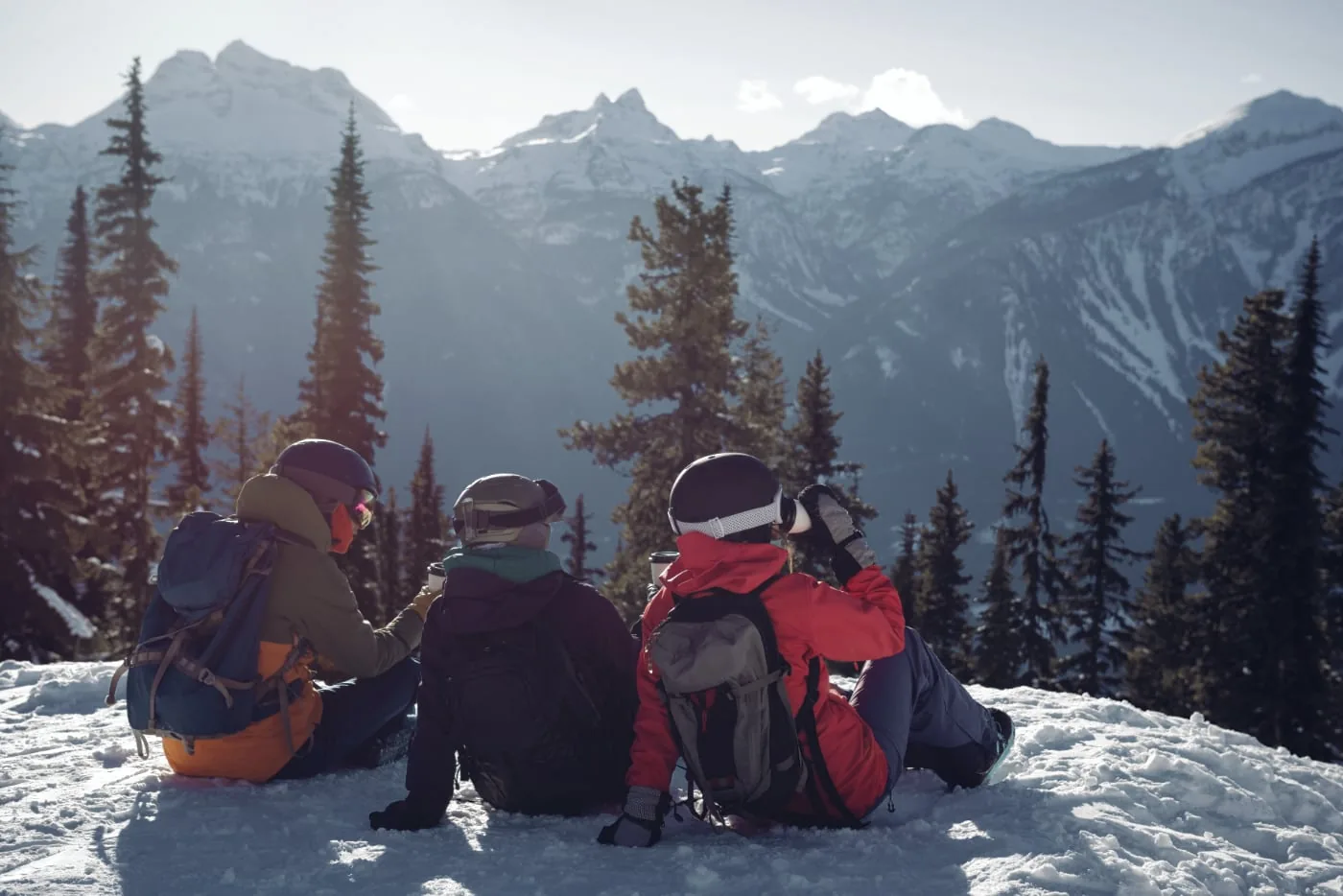 Three winter sports enthusiasts rest in the snow with snow-capped mountains in the background