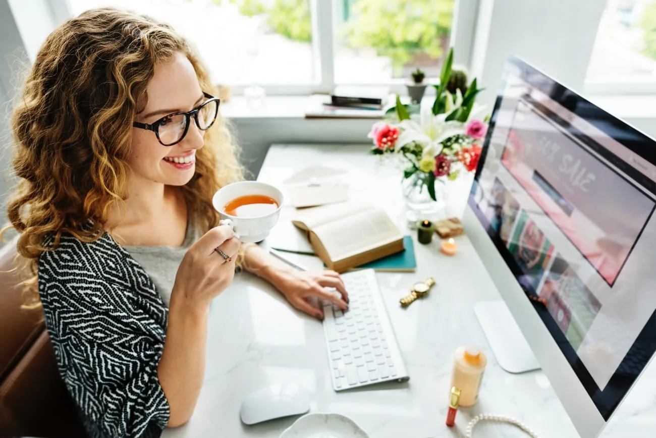 Smiling woman drinking tea at a small desk with a computer
