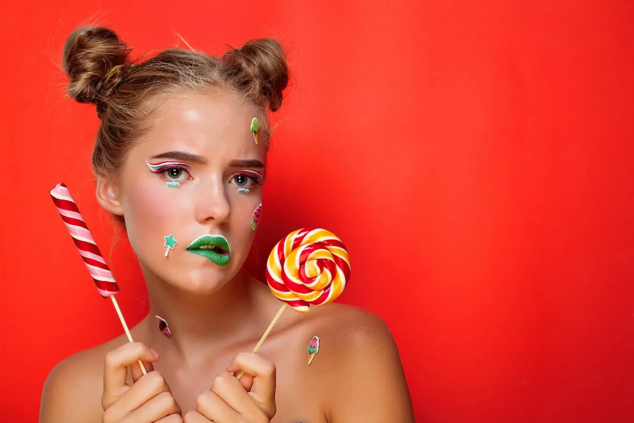 Young woman with colorful makeup and two lollipops against a red background