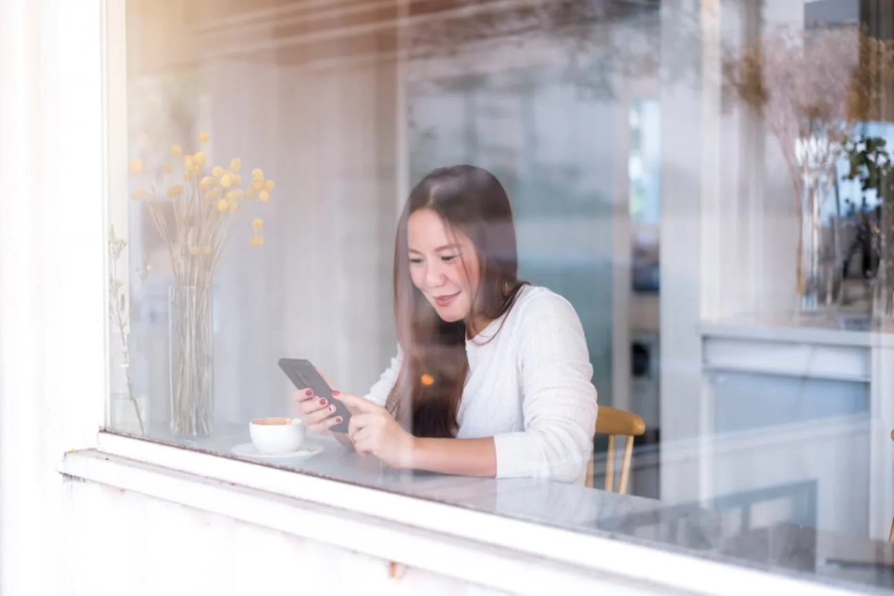 young-asian-woman-relax-using-phone-shot-through-glass-window-coffee-shop