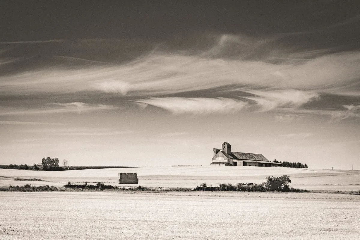 Black-and-white landscape photograph of a remote farmhouse beneath a dramatic sky