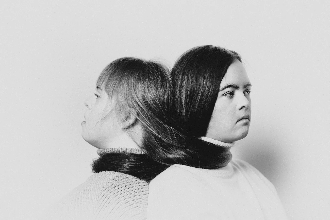 Photograph of two women with Down syndrome sitting back-to-back with their hair intertwined