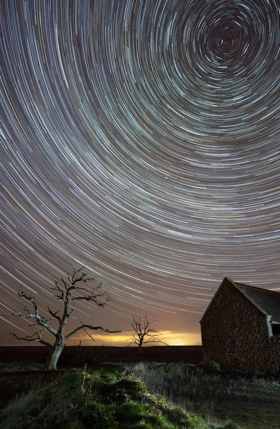 photo of a star trail above a meadow with a house and two bare trees at night