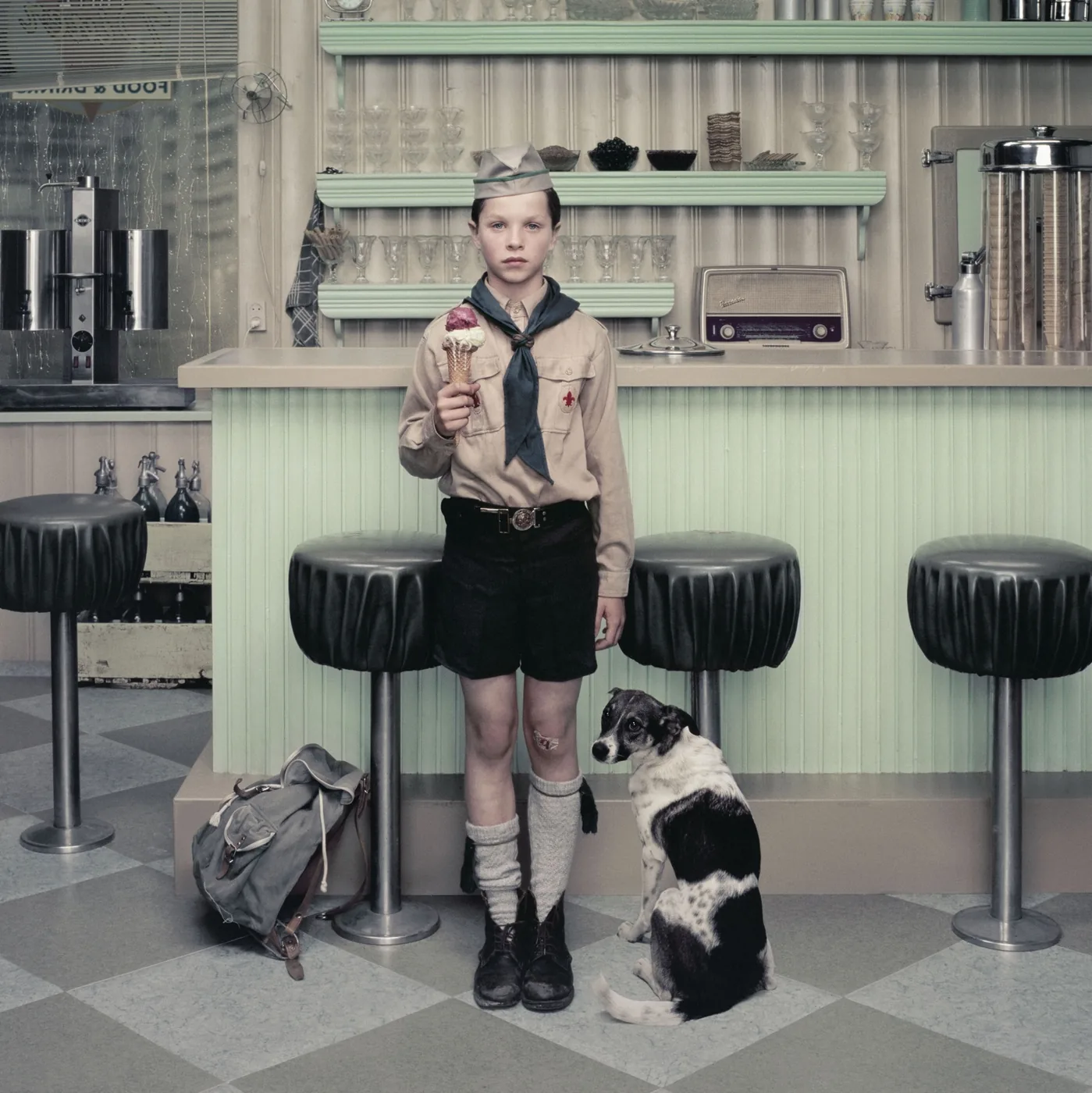 Boy around ten years old in a scout uniform inside an ice cream parlor, with a small dog beside him and his backpack on the floor
