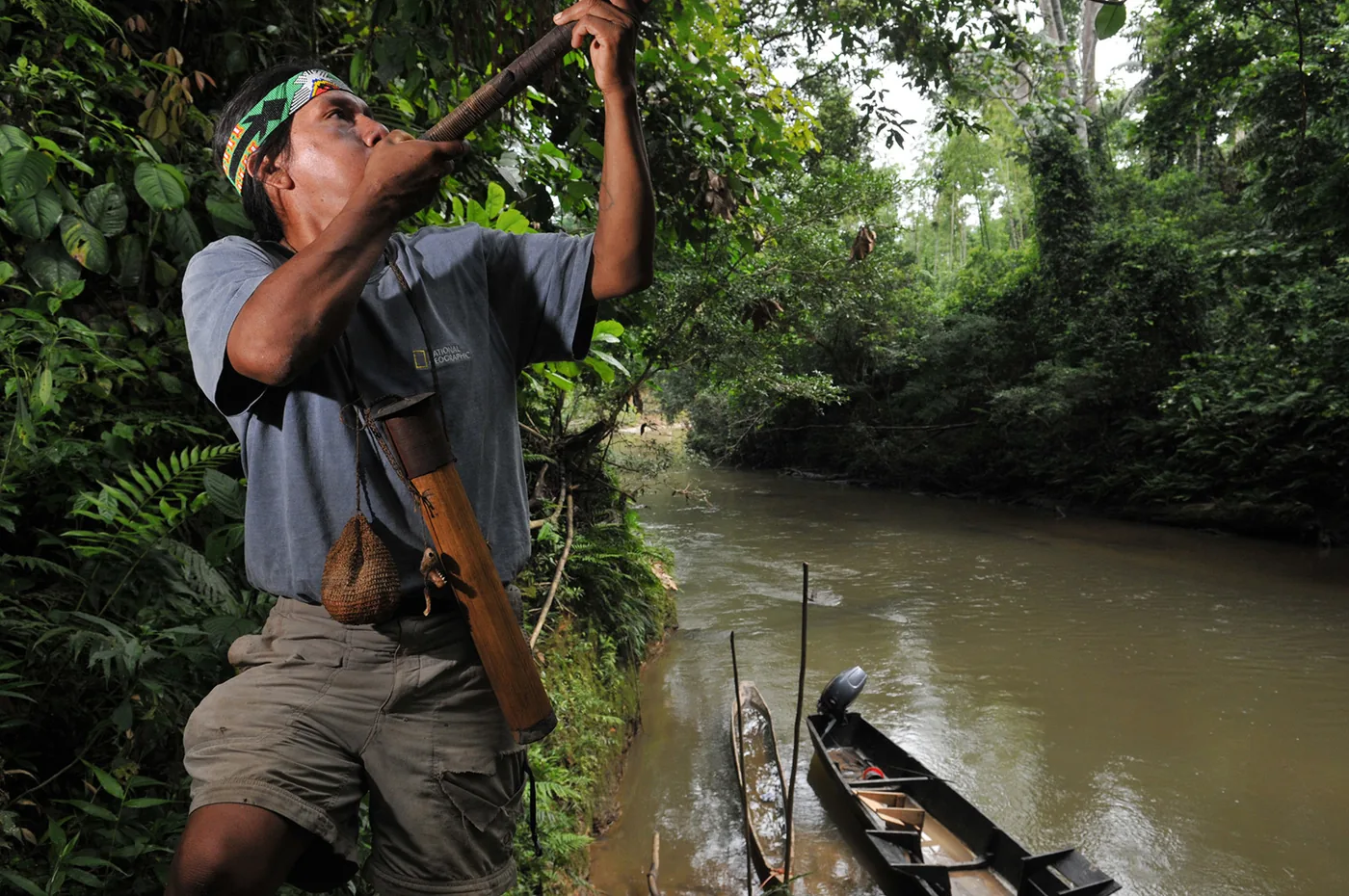 foto van een indiaan die bij een rivier met een boot in een bos blaast op een pijp die hij naar boven richt