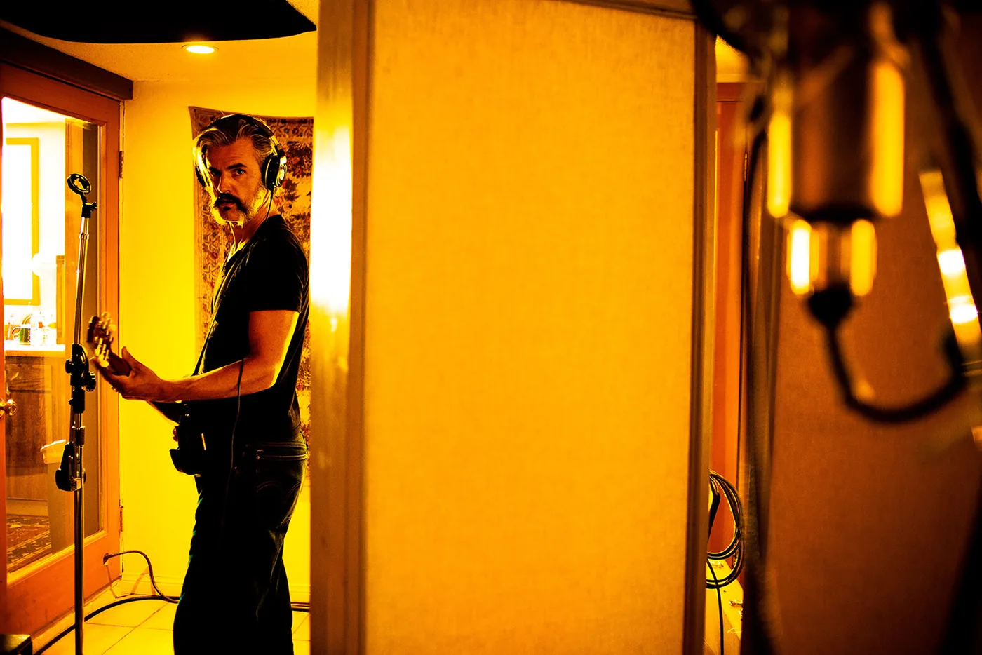 foto van een man met elektrisch gitaar en koptelefoon op in een kamer die gelig verlicht is