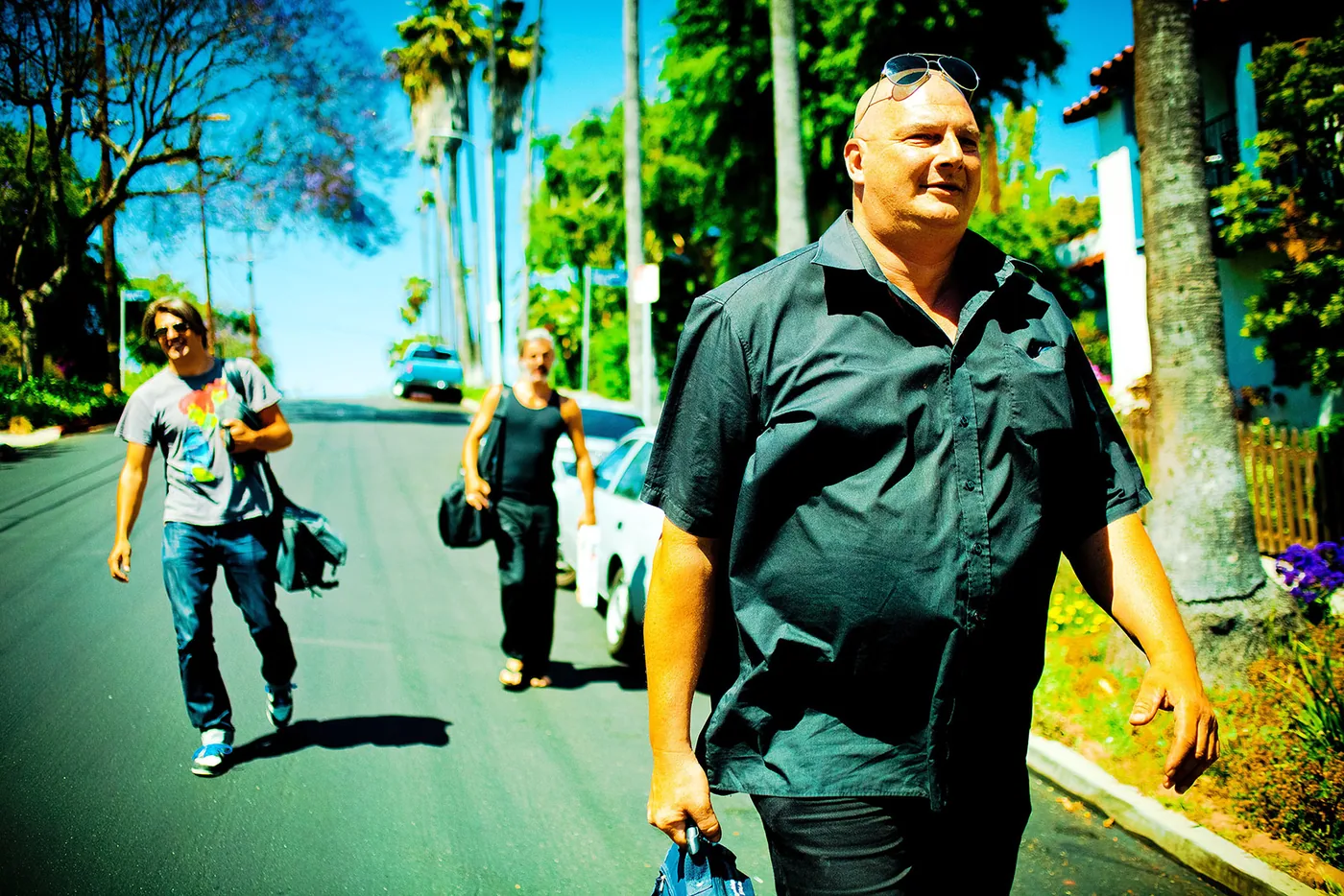 foto van drie mensen die op straat lopen in een zonnig land, een dikke man voorop en jongen links erachter en een andere man iets verder naar achter
