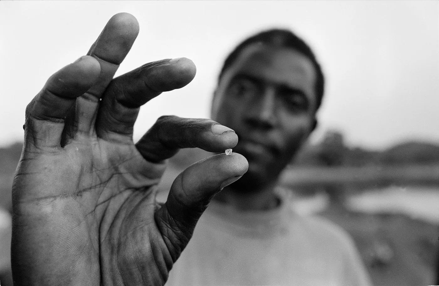 Photograph of a man’s hand holding a small diamond, with his face and the landscape faintly visible in the background