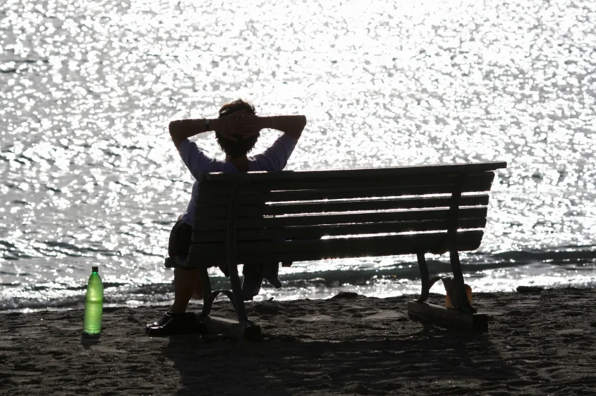 Backlit photo of a woman on a bench by the water, with reflections and a green bottle on the ground