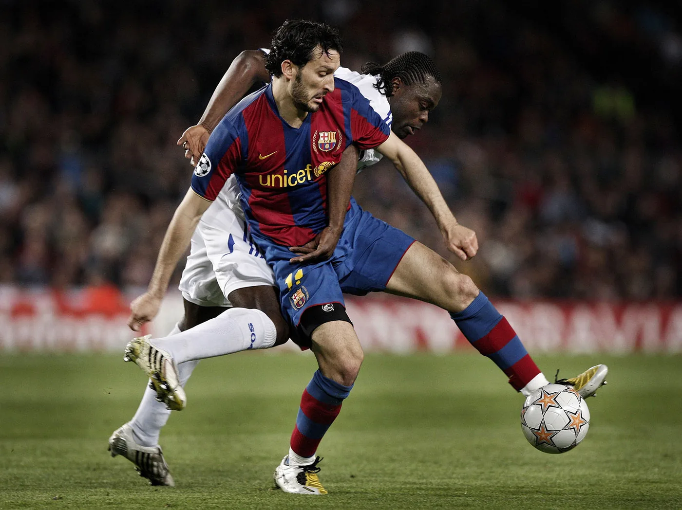 Action during a soccer match: a player in a red-and-blue uniform shields the ball as an opponent in white and blue defends, with the field and crowd in the background.