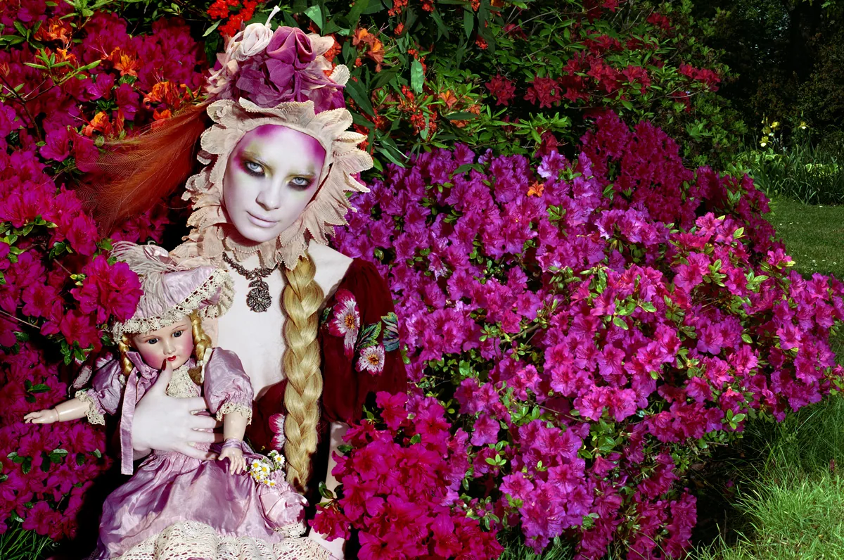 photograph by Miles Aldridge of a fairytale-like woman among flowering shrubs, with a blonde braid, a doll in her arms, and a flower hat