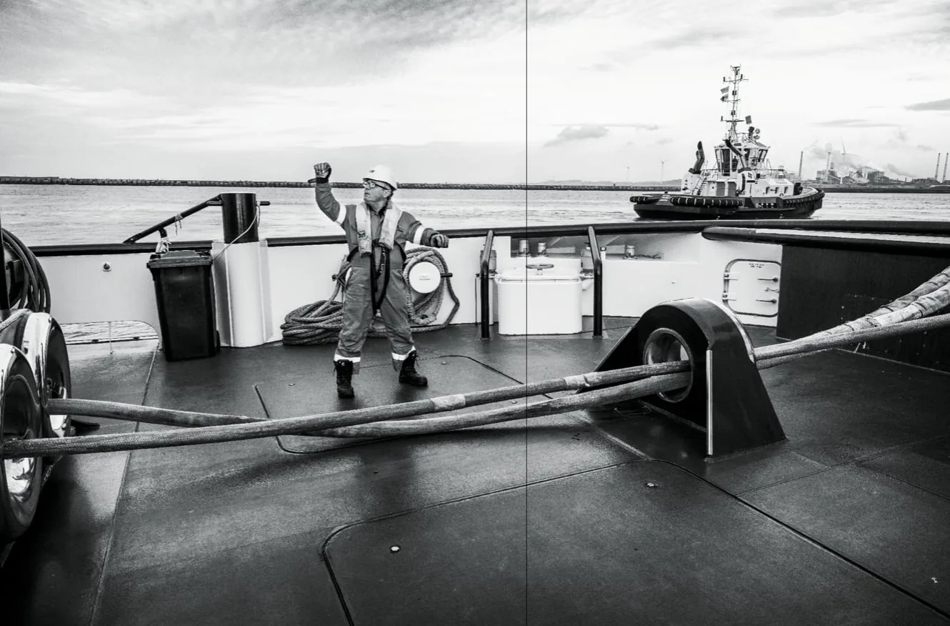 Zwart-witfoto van een persoon in werkkleding op het dek van een schip met een sleepboot op de achtergrond