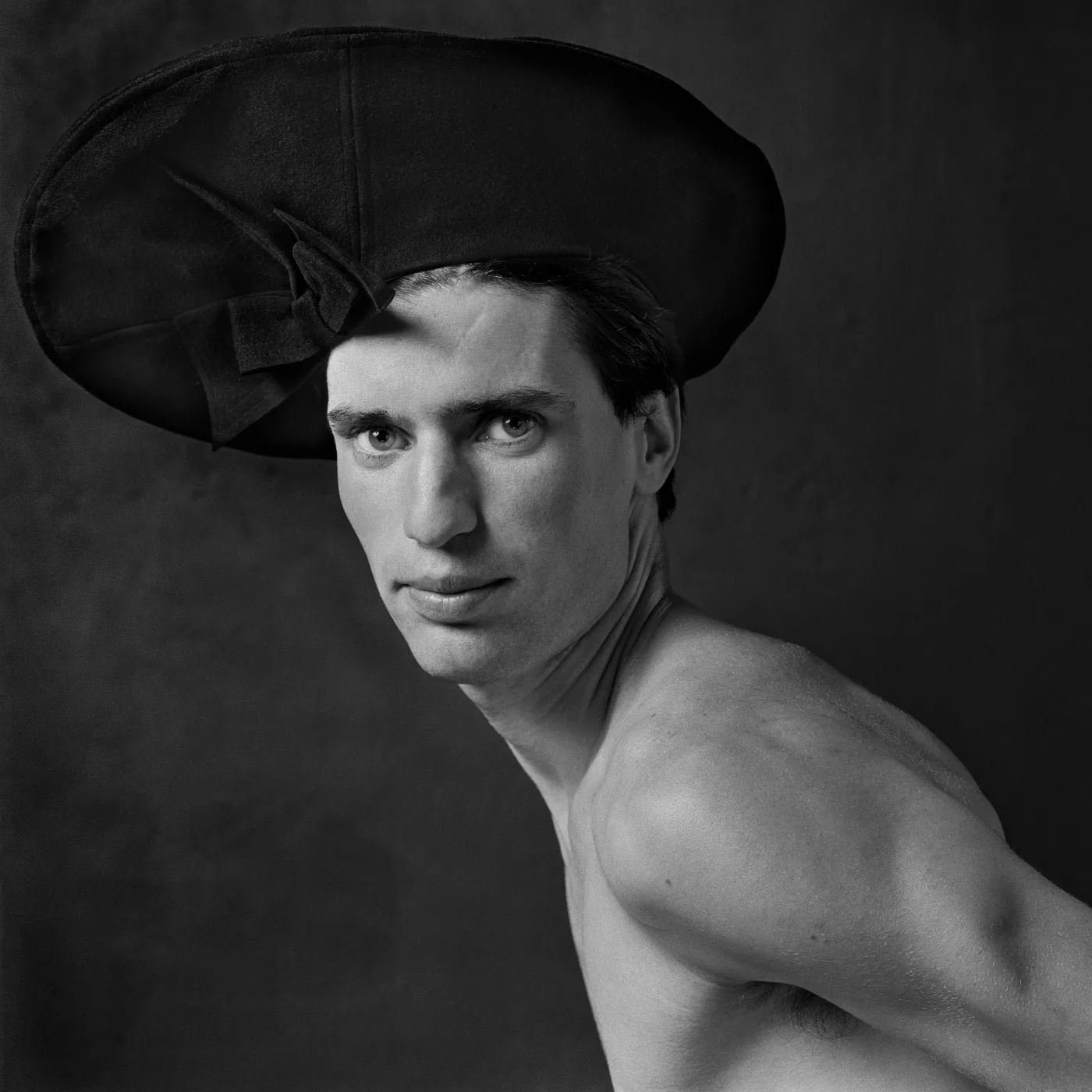 Black-and-white portrait of a half-dressed man looking over his shoulder while wearing a large women’s hat, from Erwin Olaf’s Ladies Hats series (Hennie, 1985)