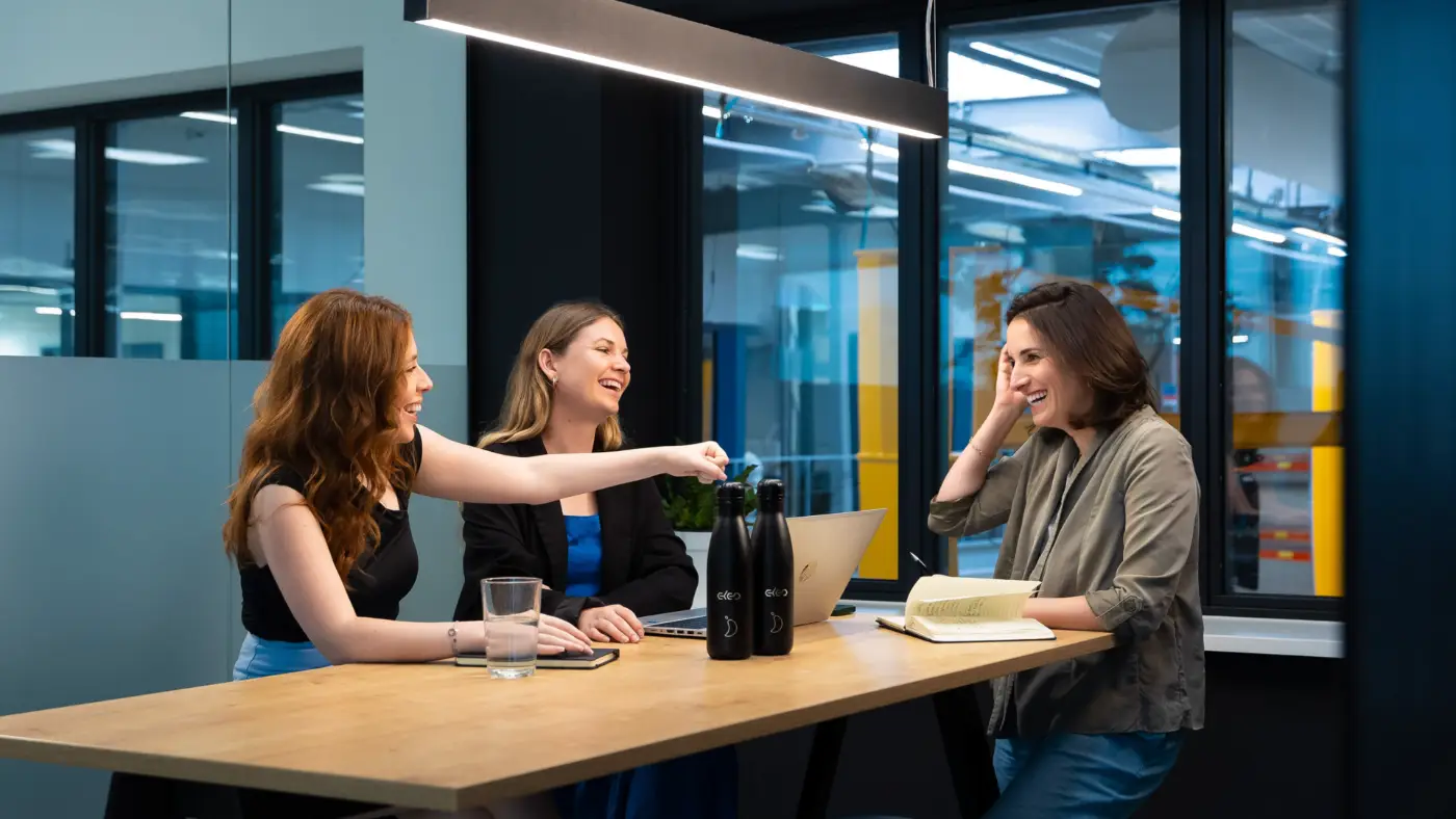 Drie lachende vrouwen in gesprek rond een tafel op kantoor