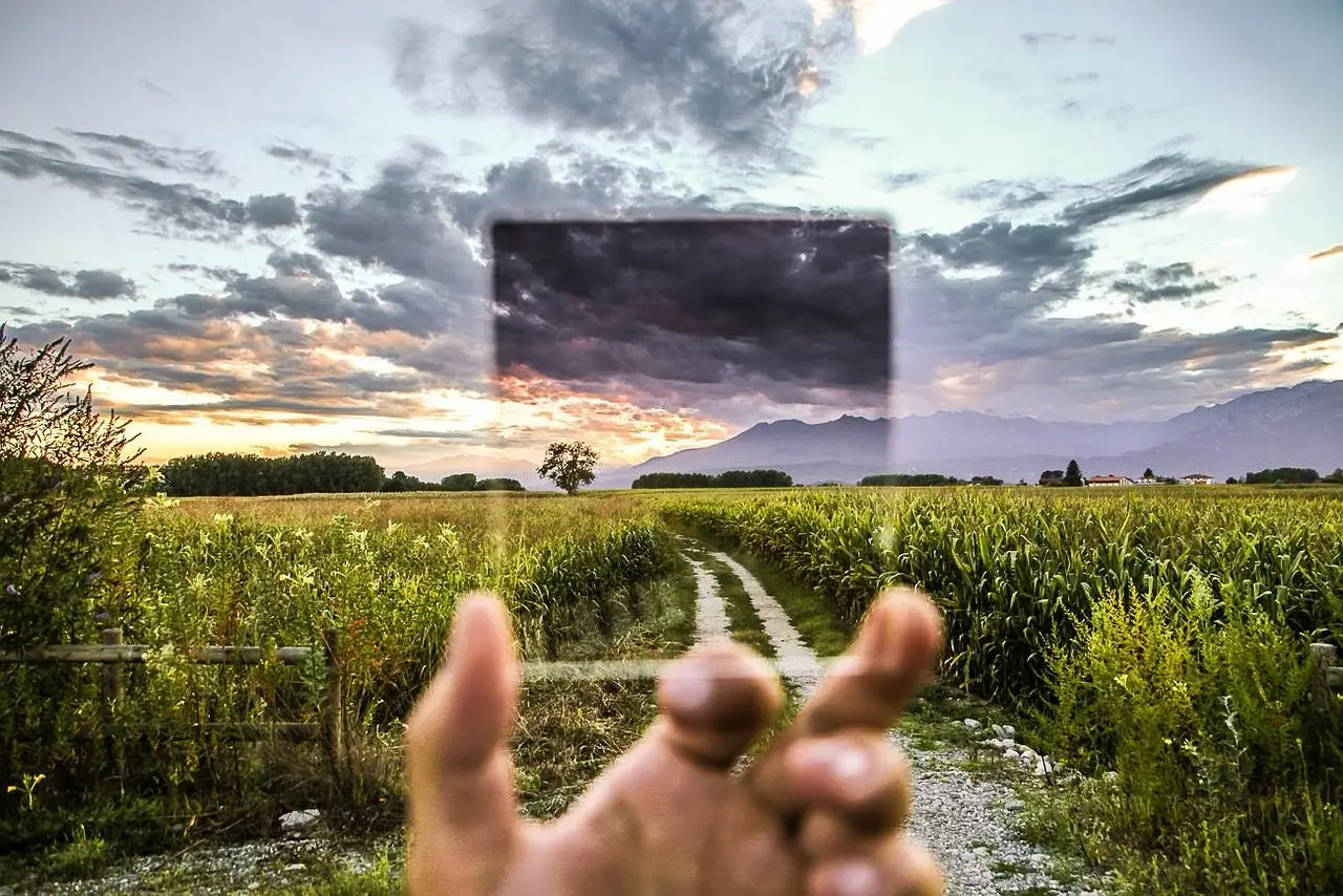 Hand houdt een polarisatiefilter voor een landschap met akkers, bergen en een bewolkte avondlucht.