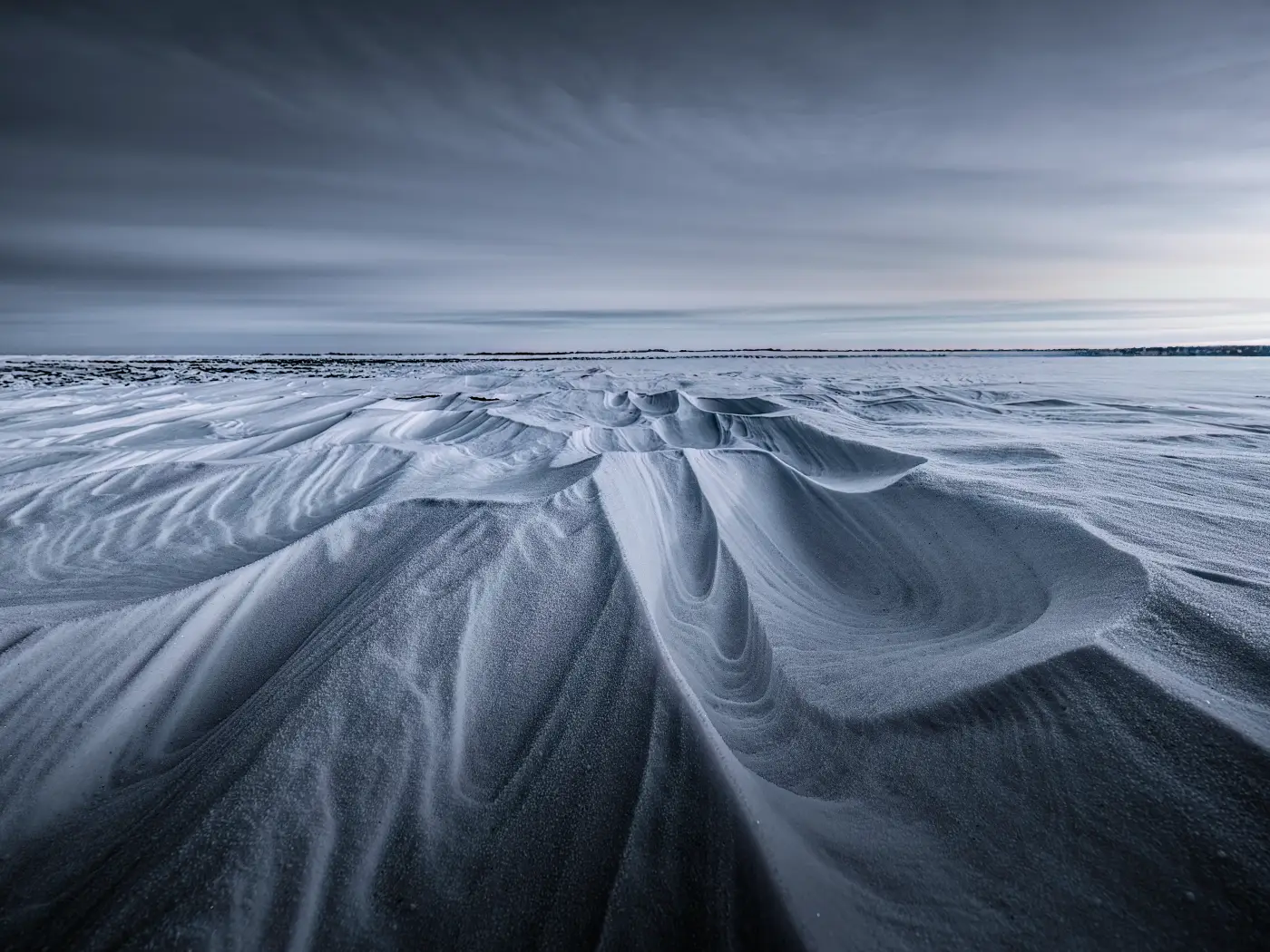 Snow-covered landscape with wind-shaped snow dunes beneath a dark, cloudy sky