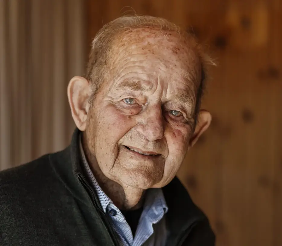 Portret van een oudere man in een donker vest en lichtblauw overhemd, gefotografeerd binnen tegen een houten achtergrond.
