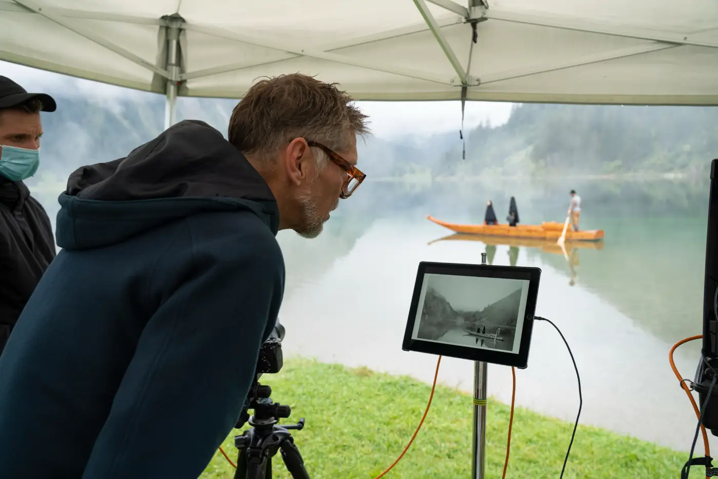 Een man bekijkt onder een tent een monitor tijdens een fotoshoot aan een meer, met een houten boot op de achtergrond.