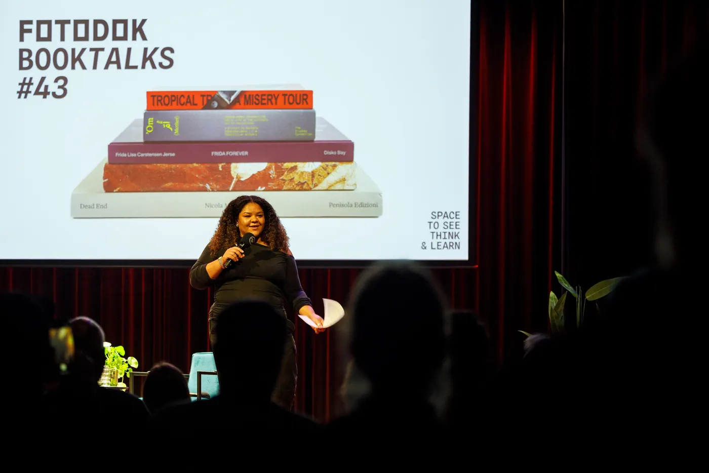 Host Giya Makondo-Wills speaks on stage during FOTODOK BOOK TALKS #43, with photobooks displayed on the screen behind her.