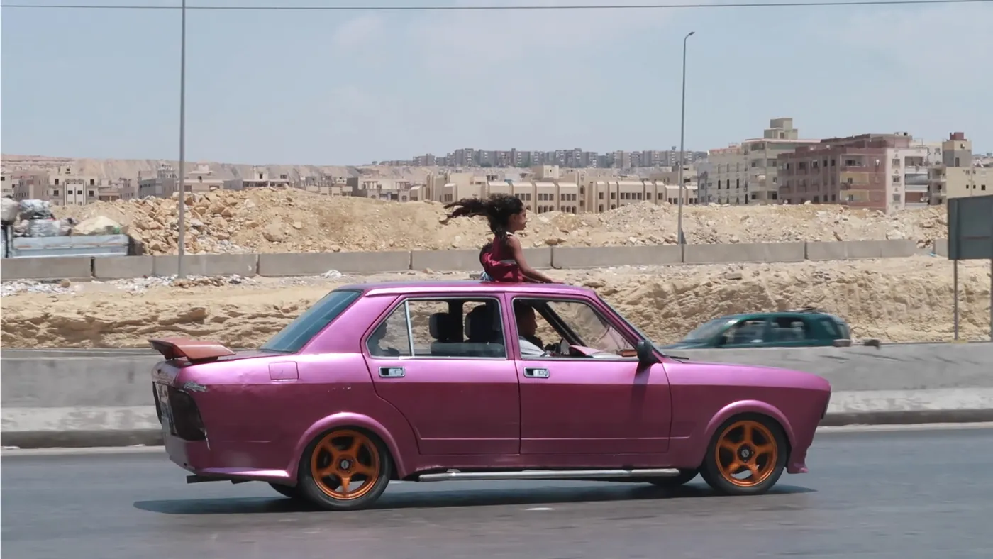 Een meisje staat door het open dak van een rijdende roze auto in Caïro, gefotografeerd door Yomna El Beyaly.