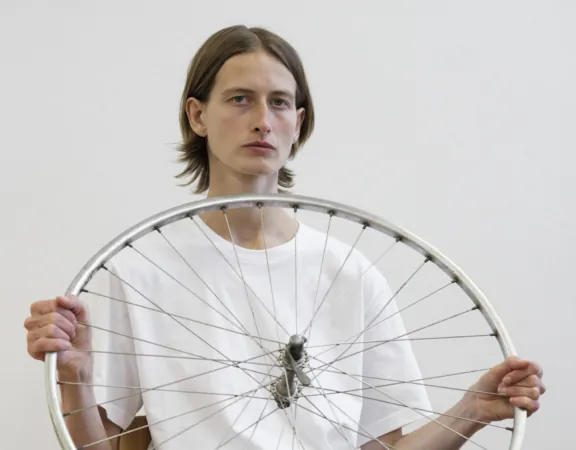 Een persoon in een wit T-shirt houdt zittend een fietswiel voor zich vast in werk van Andrzej Steinbach.