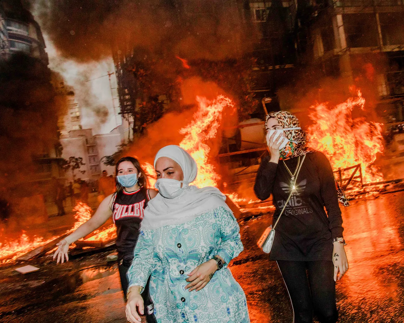 Three masked women walk past large fires in a city street, photographed by Myriam Boulos.