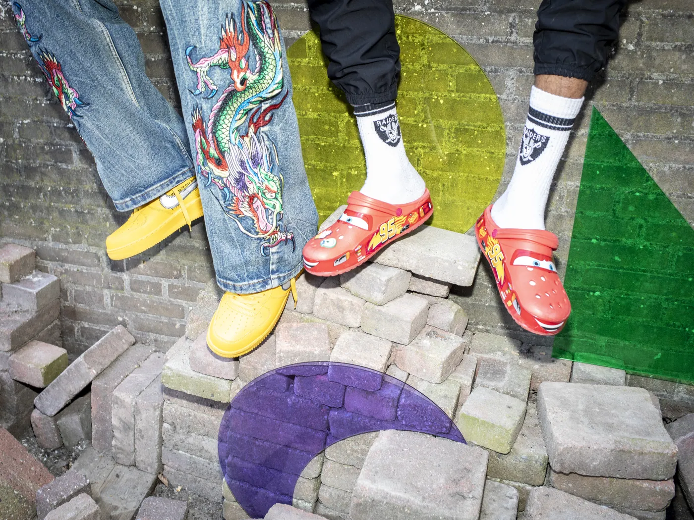 Benen met een geborduurde spijkerbroek, gele schoenen en rode klompen boven bakstenen, gecombineerd met transparante gekleurde vormen.