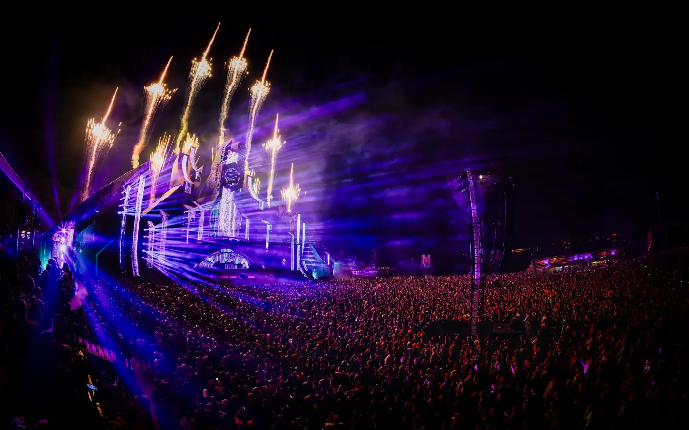 Nachtelijk festivalpodium met vuurwerk, paarse verlichting en een grote menigte in fisheye-perspectief.