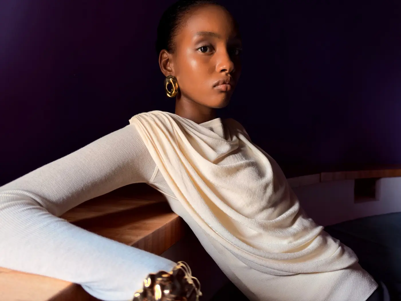Portret van een vrouw in een crèmekleurige outfit met gouden sieraden tegen een donkerpaarse achtergrond, gemaakt met de iPhone 18 Pro.