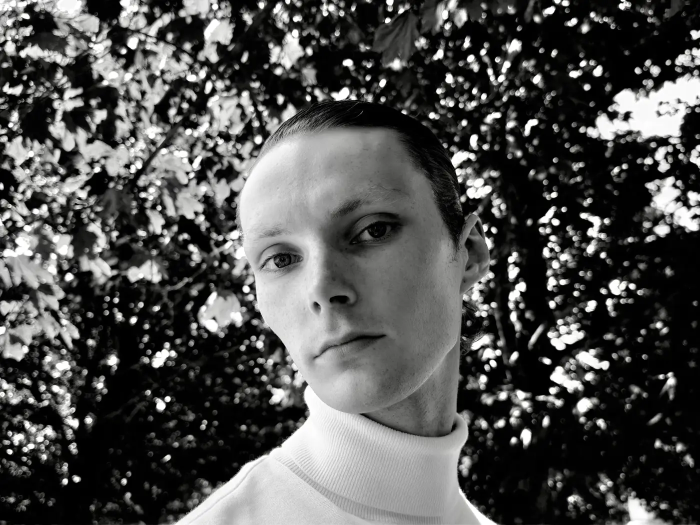 Zwart-witportret van een persoon met een lichte coltrui voor gebladerte met lichtvlekken, gemaakt met de iPhone 18 Pro.