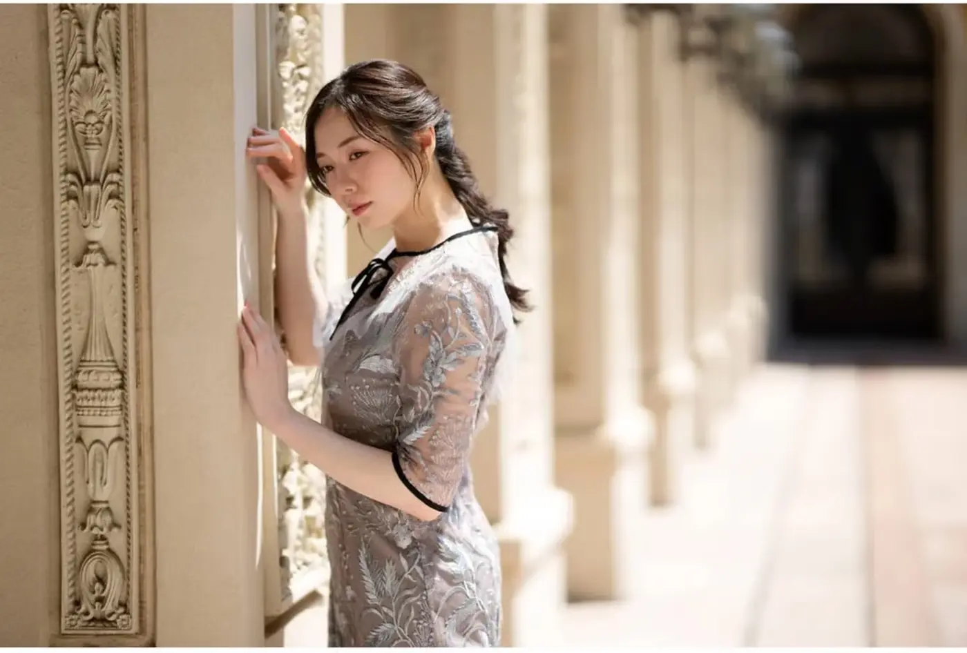 A woman leans against an ornate stone column in a gallery with a strongly blurred background.